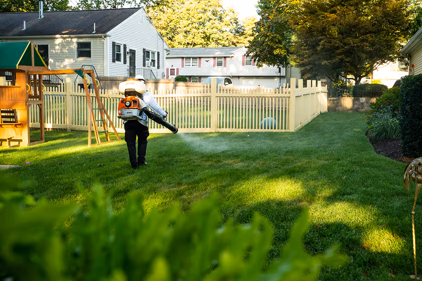 Person wearing protective gear using a leaf blower on a green lawn in a backyard with a wooden playset and white picket fence.