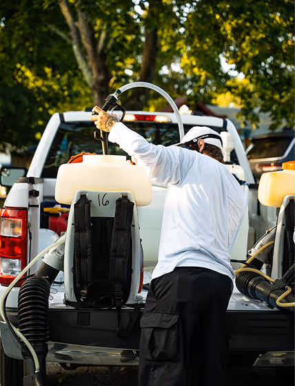 Person in white shirt preparing backpack sprayer in the bed of a pickup truck outdoors with trees in the background.