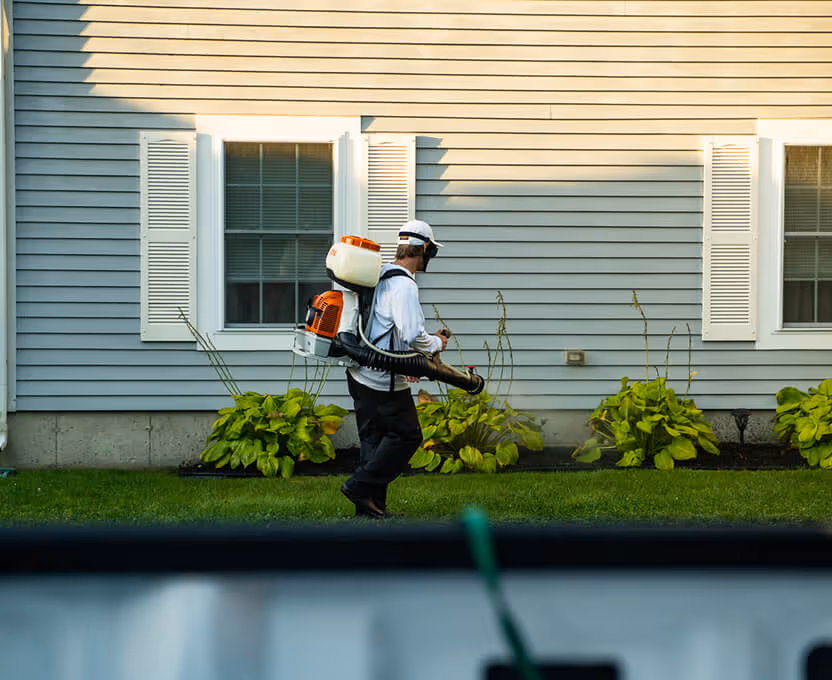 Person wearing protective gear and carrying a backpack leaf blower in a residential lawn garden.
