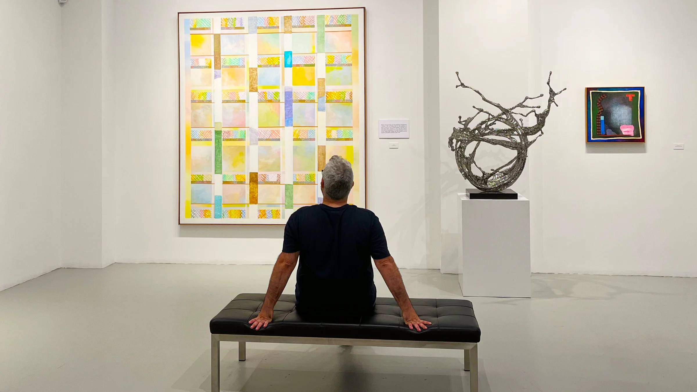 Person sitting on a bench in an art gallery viewing a large colorful abstract painting, a metal sculpture on a pedestal, and a smaller abstract painting on the wall.
