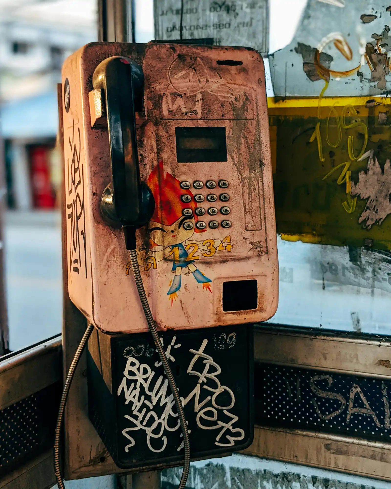 Worn public payphone with graffiti and cartoon decals inside a glass booth.