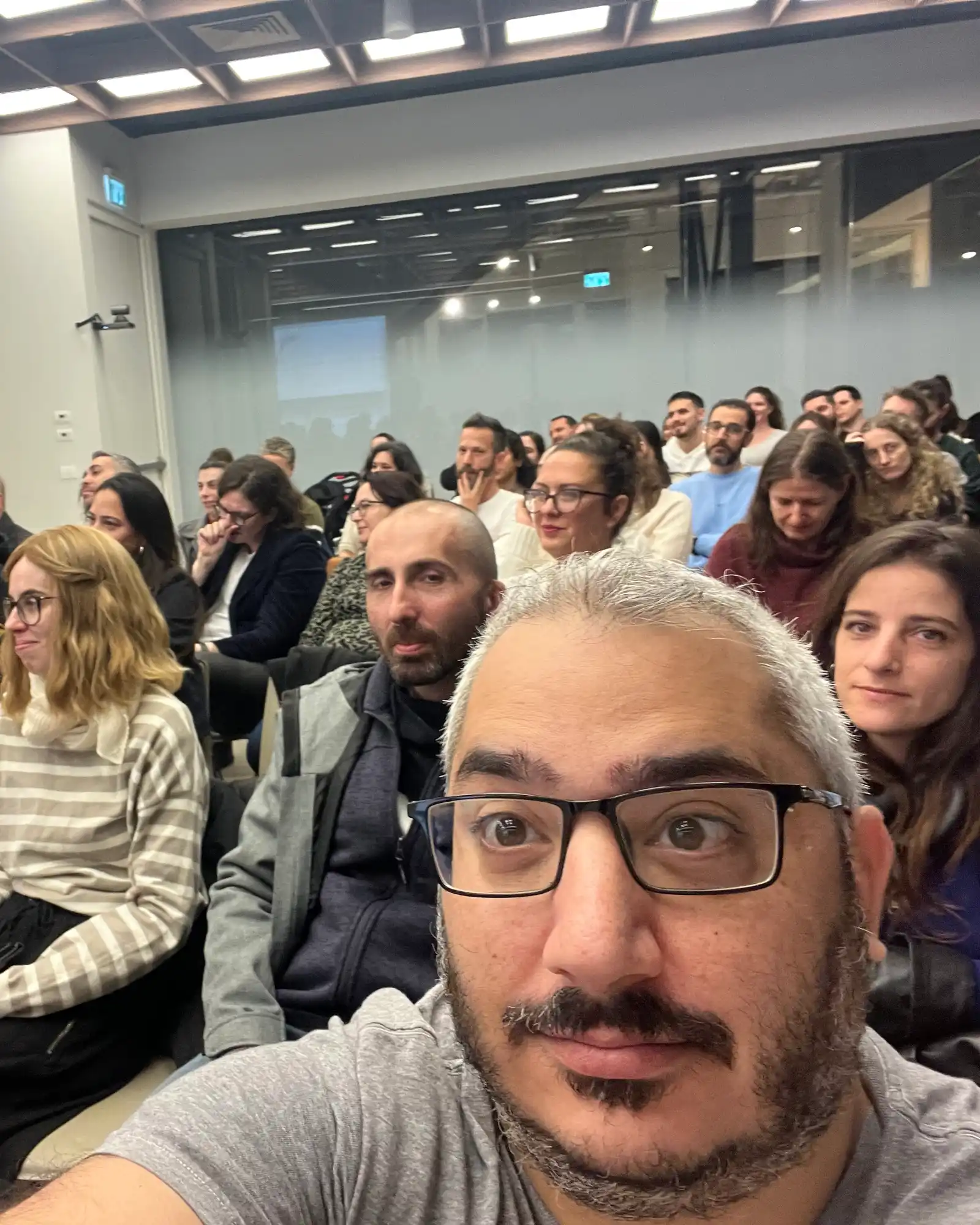 Man with glasses and gray hair taking a selfie in front of a room full of seated people.