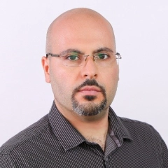 Portrait of a bald man with a beard and glasses wearing a dark striped shirt against a plain white background.