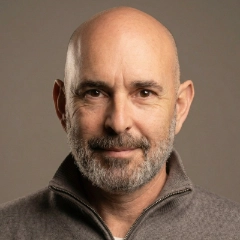 Portrait of a bald man with a beard and glasses wearing a dark striped shirt against a plain white background.