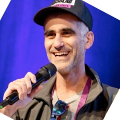 Man wearing a cap and holding a microphone, smiling against a purple background.