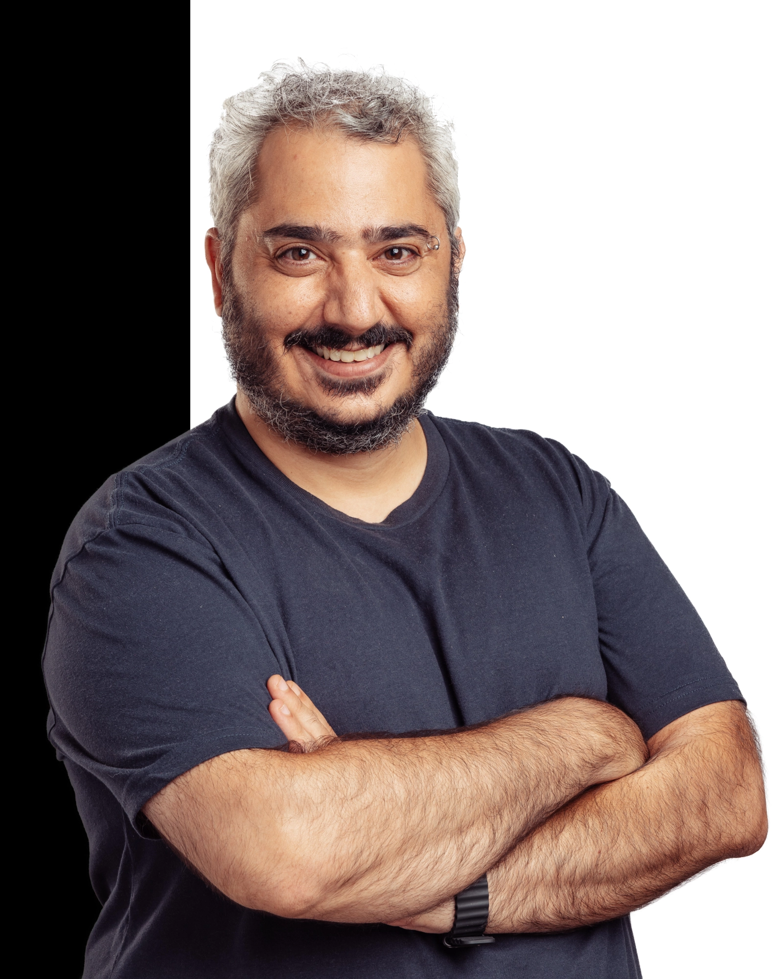 Smiling man with gray hair and beard wearing a dark blue t-shirt and a black wristwatch standing with his arms crossed against a white background.