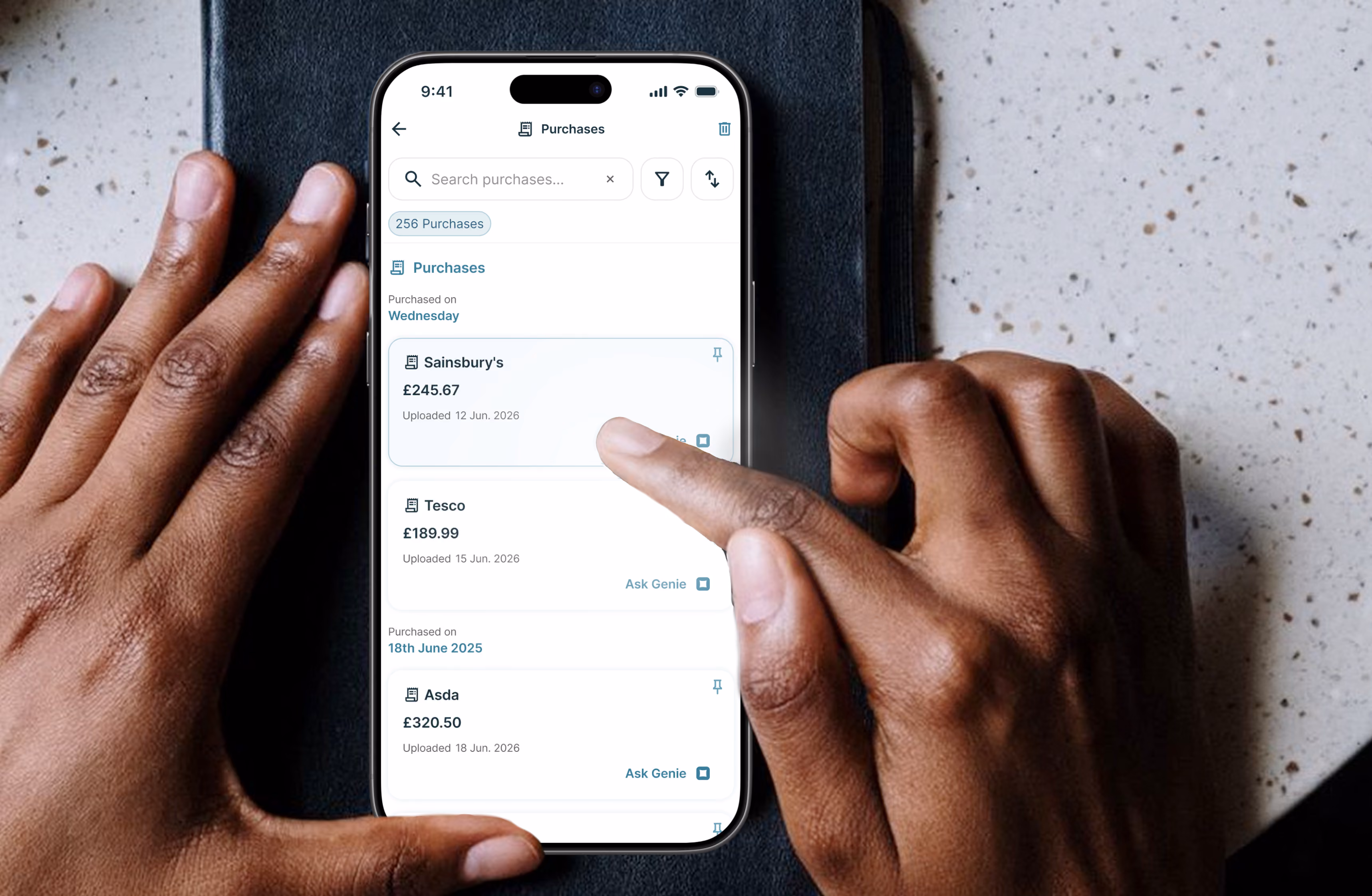 Hands holding a phone showing a purchases app with entries for Sainsbury's, Tesco, and Asda with amounts.