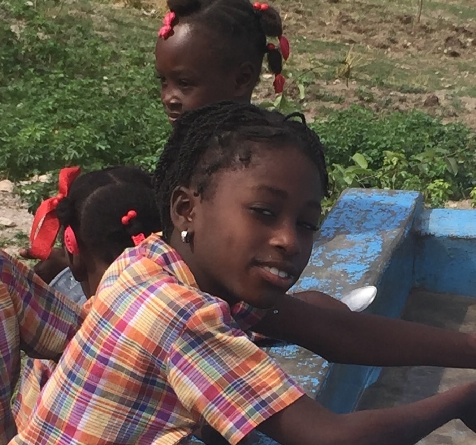 Three girls with braided hair and red hair accessories wearing plaid shirts near a blue water trough outdoors.