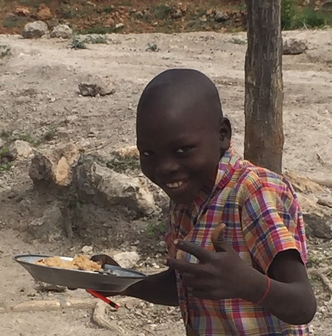 Smiling boy in a colorful checkered shirt holding a metal plate with food outdoors.