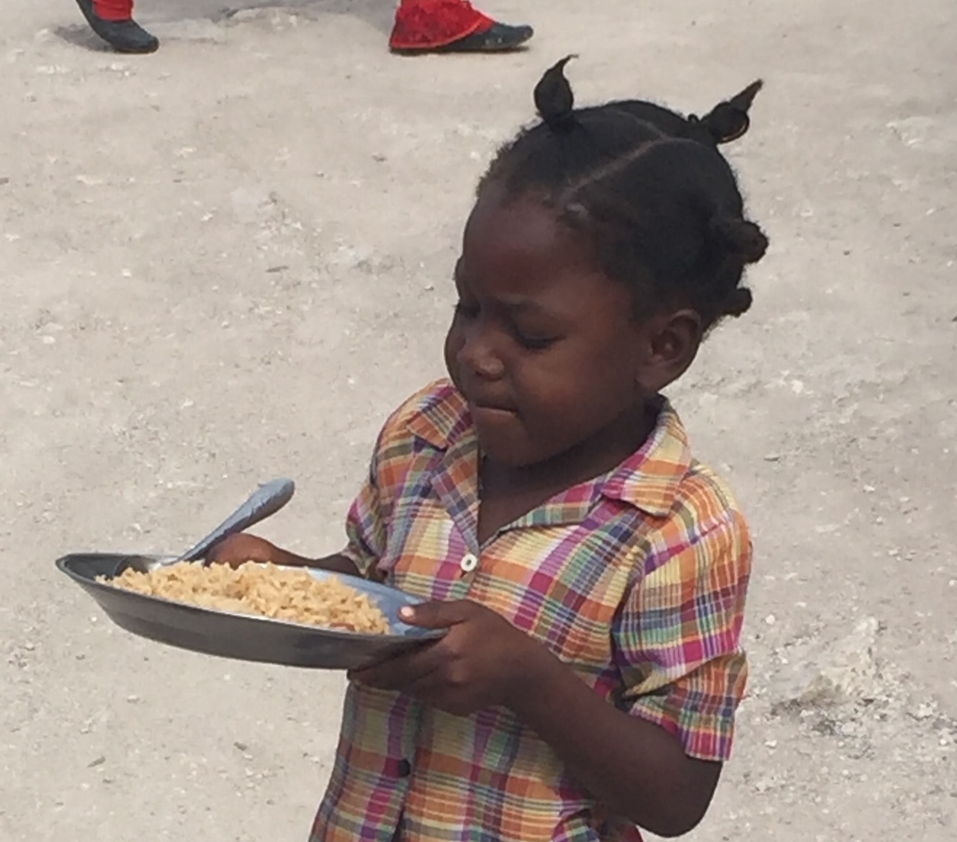 Young child in a colorful plaid shirt holding a plate of rice with a spoon outside on a dirt ground.