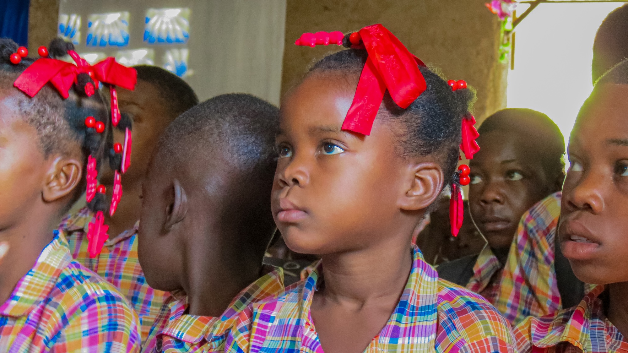 Young children in Bayonnais Haiti in colorful plaid uniforms sitting closely, with girls wearing red hair ribbons and beads, attentively looking forward indoors.
