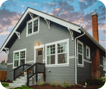 Gray wooden house with white trim, a small front porch, and a brick chimney under a blue sky with clouds.