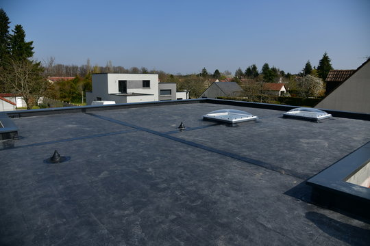 Flat black rooftop with two white skylights and ventilation pipes, suburban houses and trees in the background under a clear blue sky.