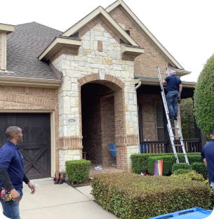 Two men working outside a brick and stone house, one on a ladder near the roof, the other standing on the driveway.