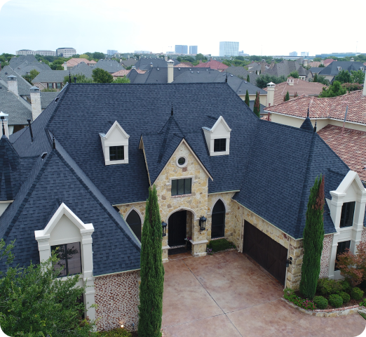 Stone and brick house with steep dark gray roof and two tall cypress trees at the front driveway.