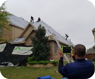 Three workers on a roof installing or repairing shingles while another person in the yard uses a tablet to monitor or document the work.