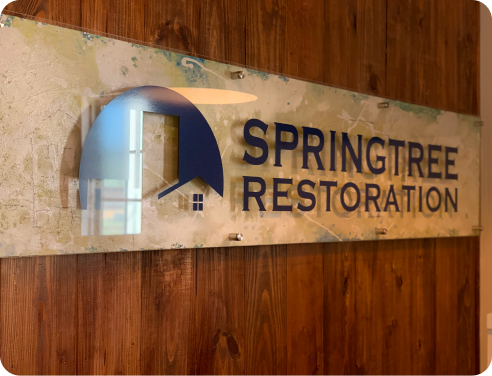 Wall-mounted translucent sign with dark blue Springtree Restoration logo and text on a wooden wall.