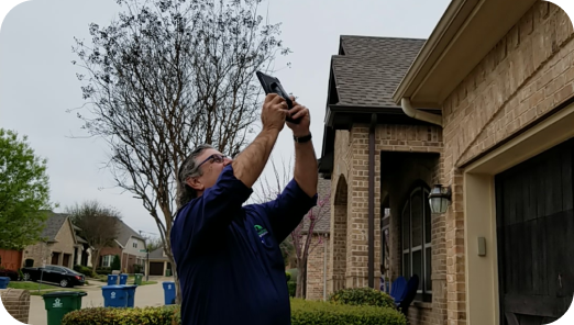 Man in navy shirt using a tool to inspect or repair the gutter of a brick house on a cloudy day.