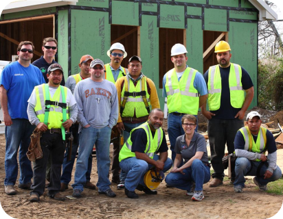 Group of construction workers wearing safety vests and helmets posing at a building site in front of a partially constructed structure.