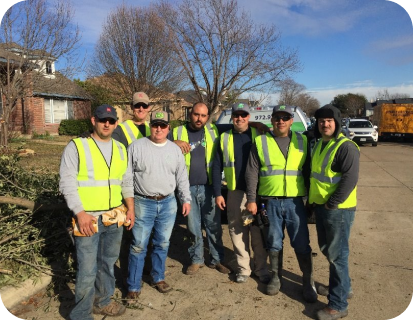 Seven men wearing yellow safety vests standing on a street in front of a house and vehicles on a sunny day.