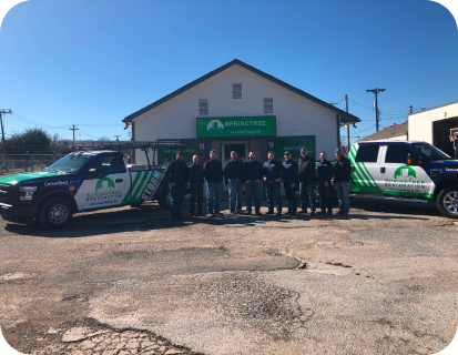 Group of twelve people standing between two branded service trucks in front of a Springtree lawn care building under a clear blue sky.