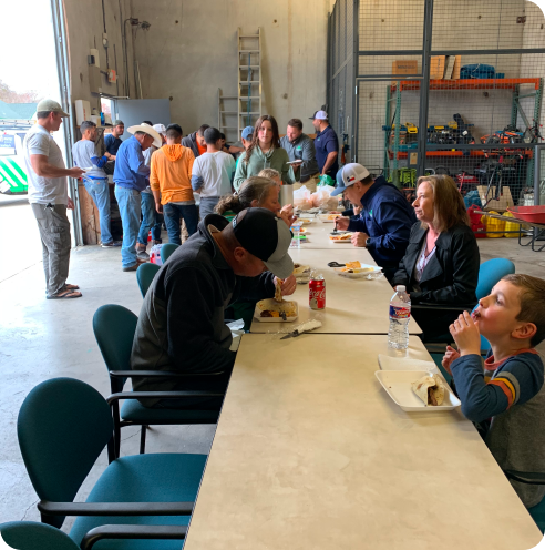 People sitting and eating at a long table inside a warehouse, while others stand in line near an open garage door.