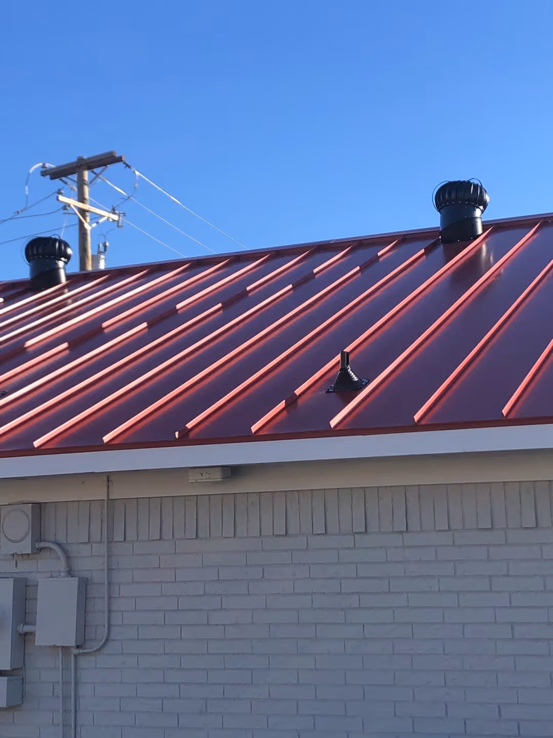 Red metal roof with two black turbine vents and a gray brick wall beneath under a clear blue sky.