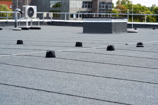 Flat rooftop with black safety anchor points and thin cables, air conditioning unit, and surrounding urban buildings.