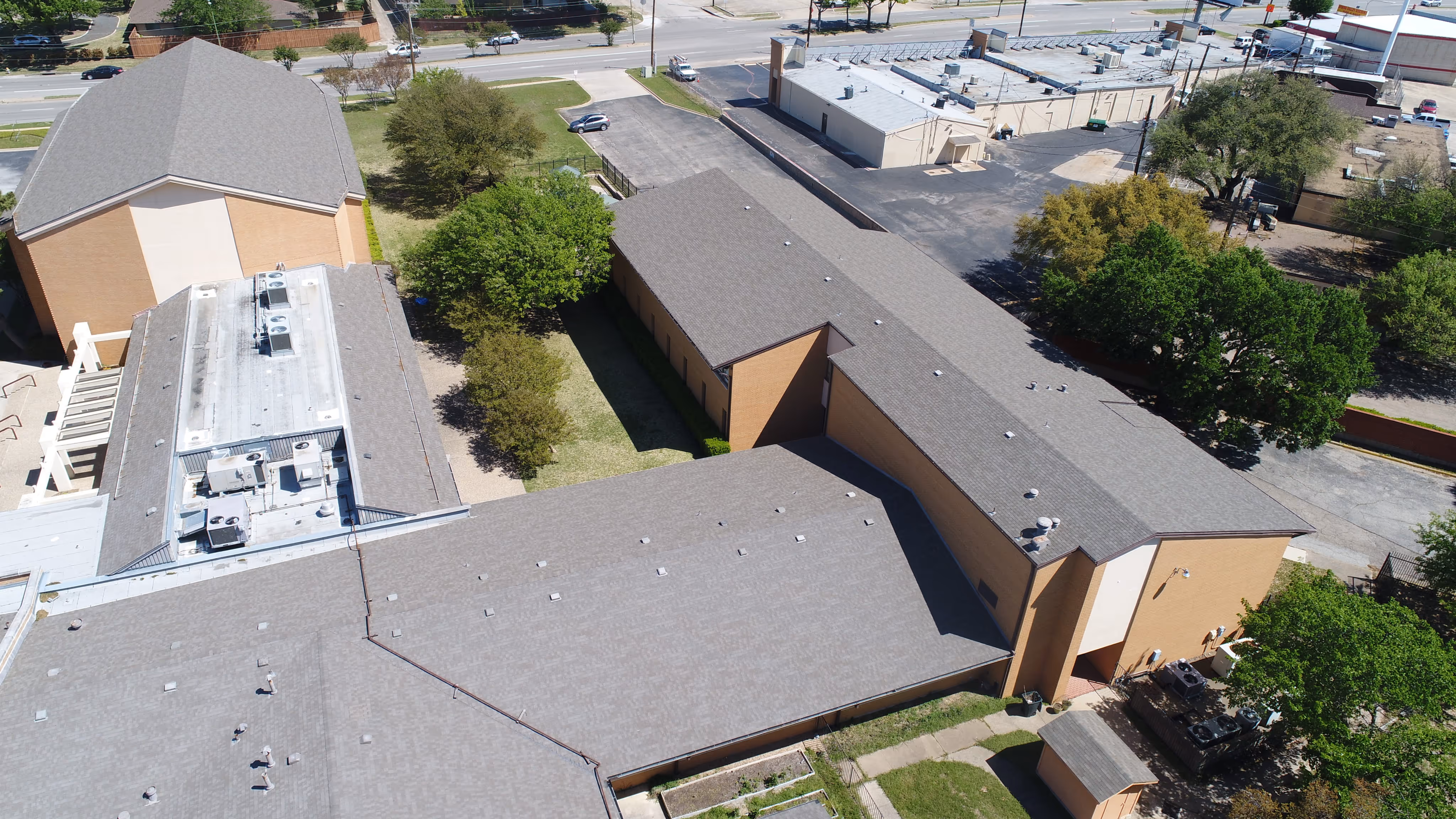 Aerial view of large beige and gray buildings with flat and pitched roofs surrounded by trees and parking areas.