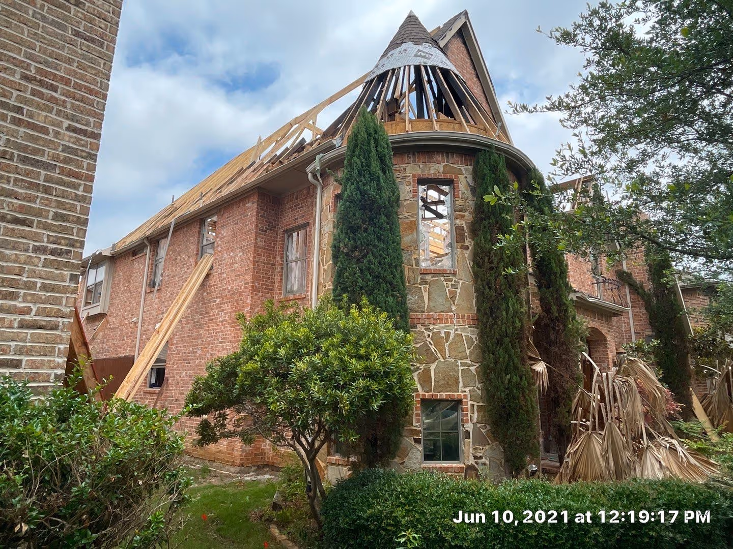 Two-story brick and stone house with partially removed roof under cloudy sky, surrounded by green shrubs and trees.