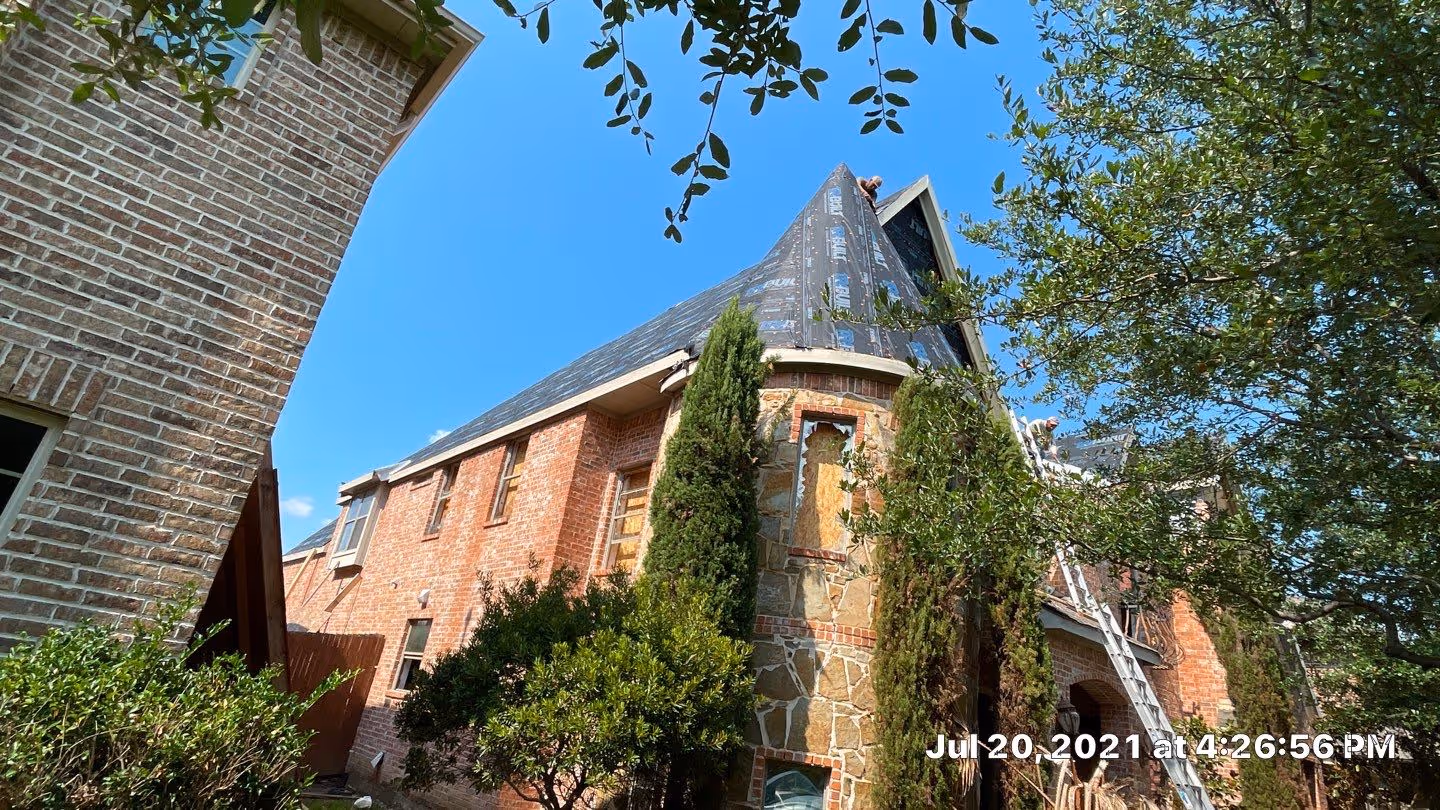 Two-story brick house with stone turret under roof repair with workers on ladders, surrounded by trees and bushes under a clear blue sky.