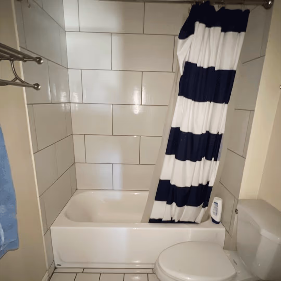 Bathroom with white tiled walls and floor, a white bathtub with a navy and white striped shower curtain, and a white toilet.