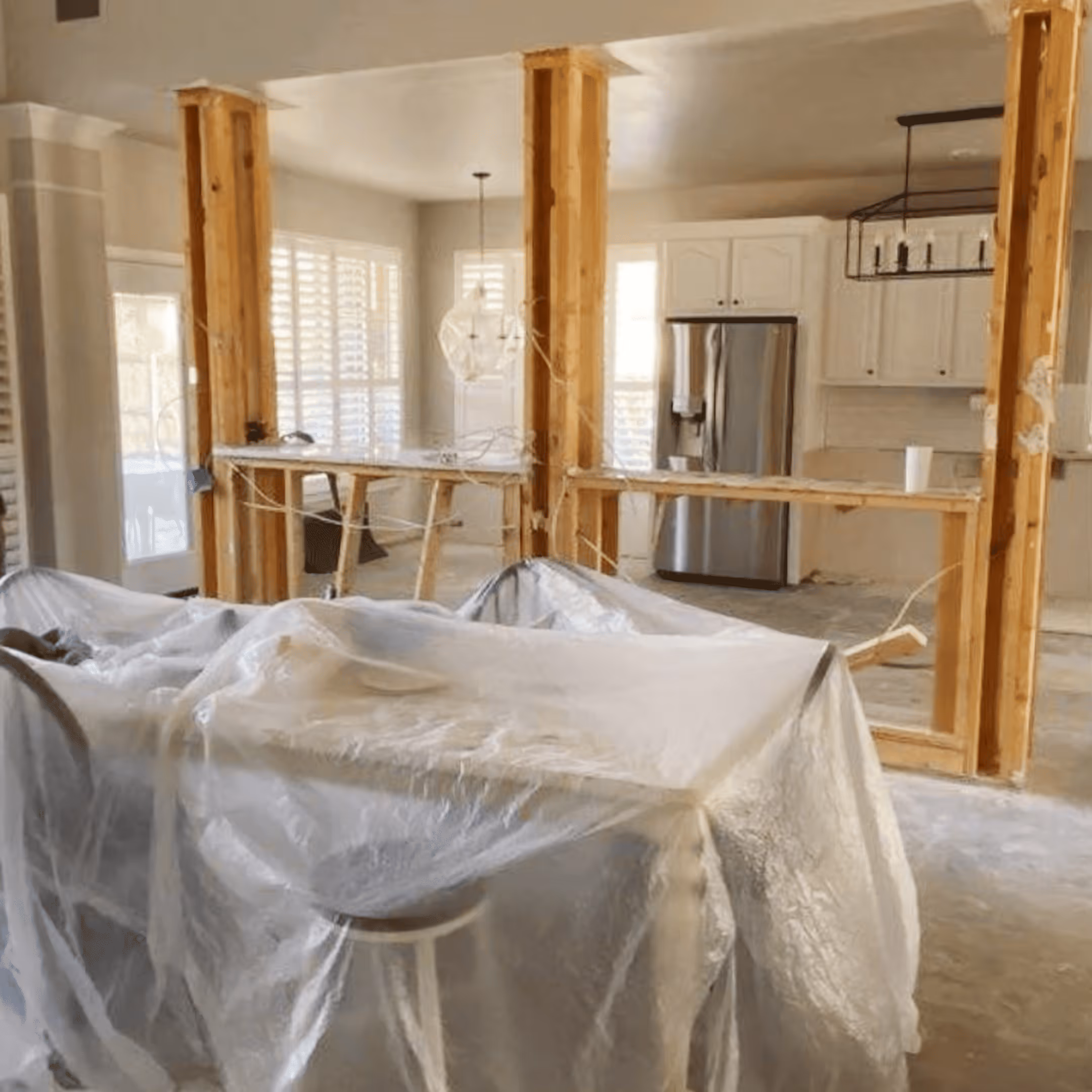 Kitchen under renovation with exposed wooden studs, plastic-covered furniture, and a stainless steel refrigerator.