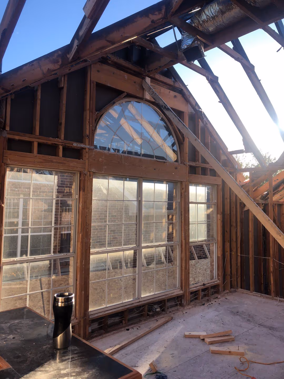 Interior view of a house under construction showing exposed wooden framing, large windows with a decorative semicircular window at the top, and construction materials on the floor.