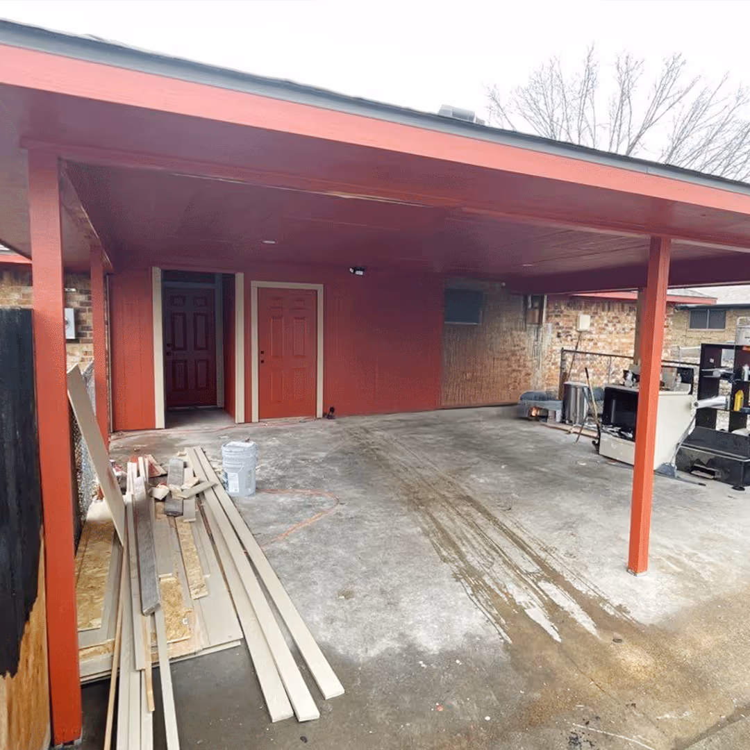 Covered concrete patio with red pillars and walls, construction lumber on the left, and some appliances and tools on the right.