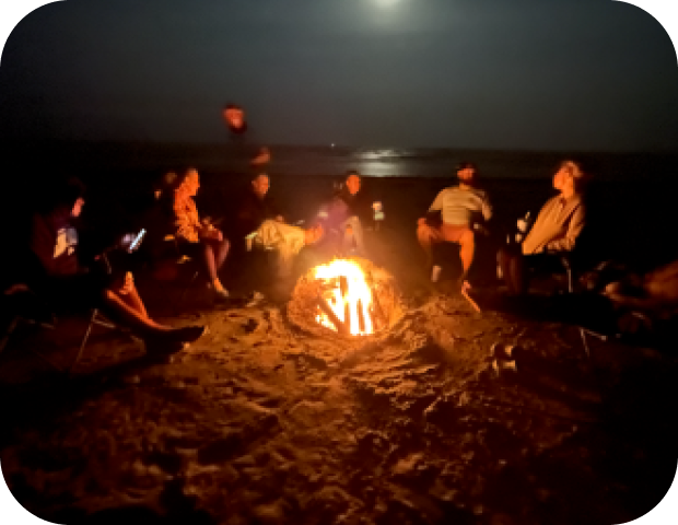 Group of people sitting around a bright campfire on a beach at night with the moon shining over the water.
