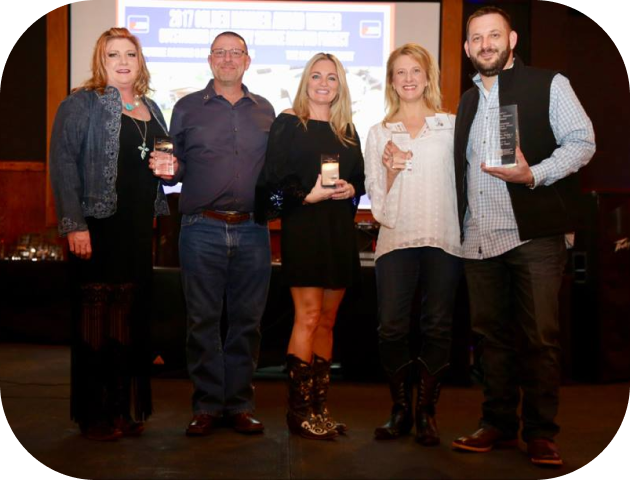 Group of five adults standing on stage holding awards, smiling at the camera in a celebratory setting.