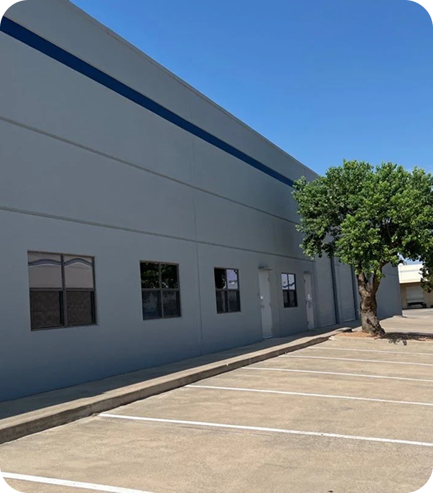 Empty parking lot beside a gray industrial building with four windows, two doors, and a leafy tree under a clear blue sky.
