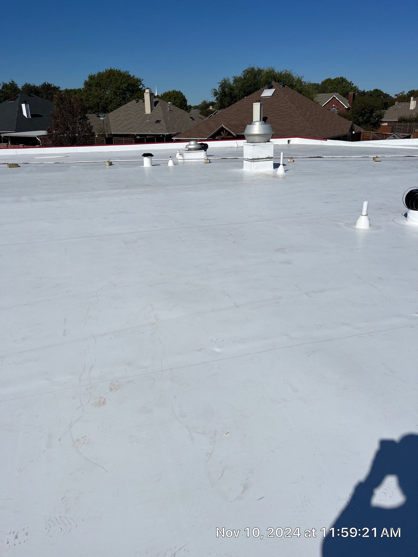 Flat white rooftop with multiple vents and chimneys, neighboring houses and trees in the background under a clear blue sky.