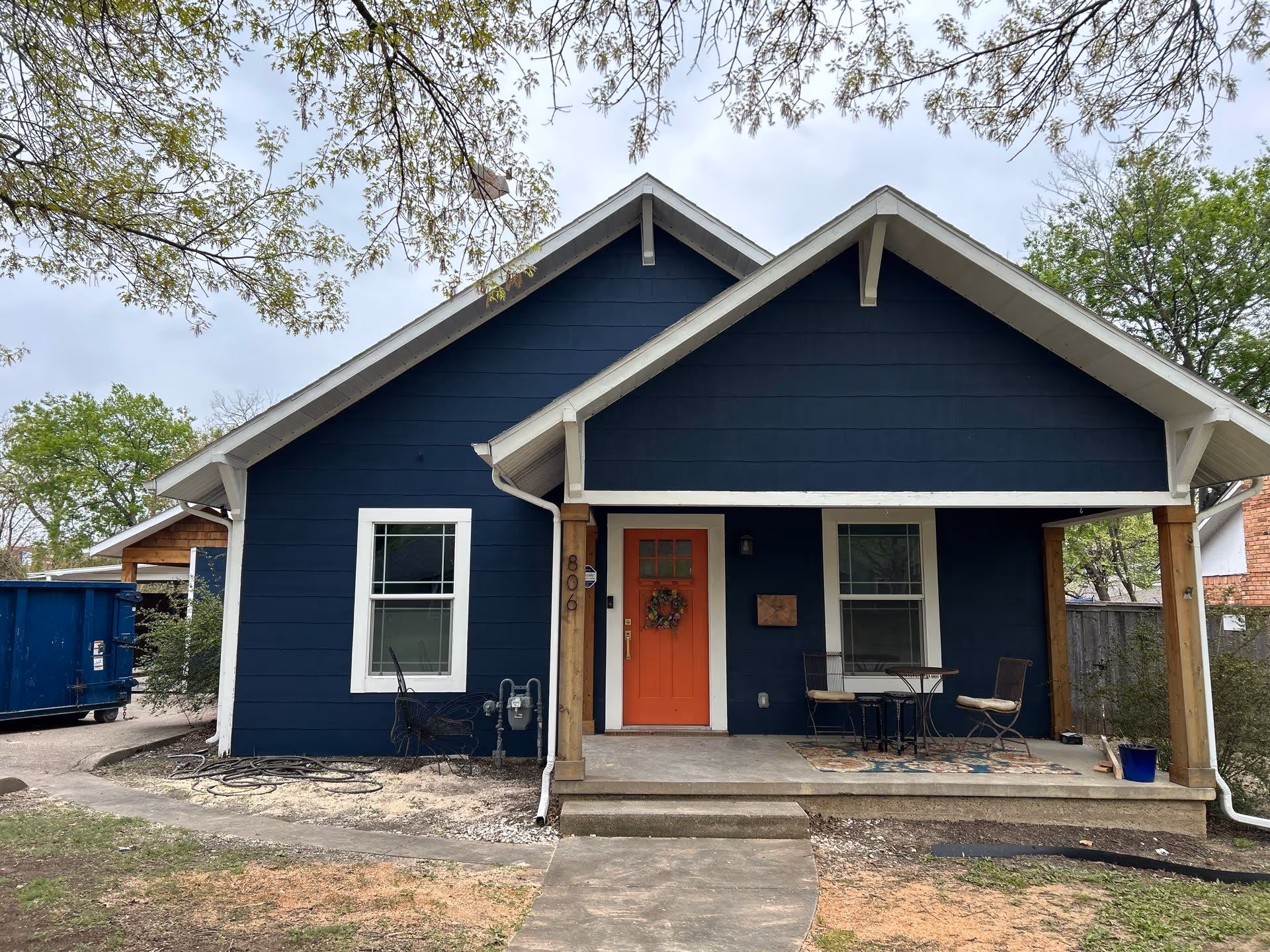 Small blue house with white trim and orange front door with wreath, a front porch with two chairs and a table, and a large tree branch overhead.