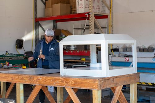 Worker in a workshop wearing a cap and jacket standing next to a wooden table with a large white metal frame structure on it.