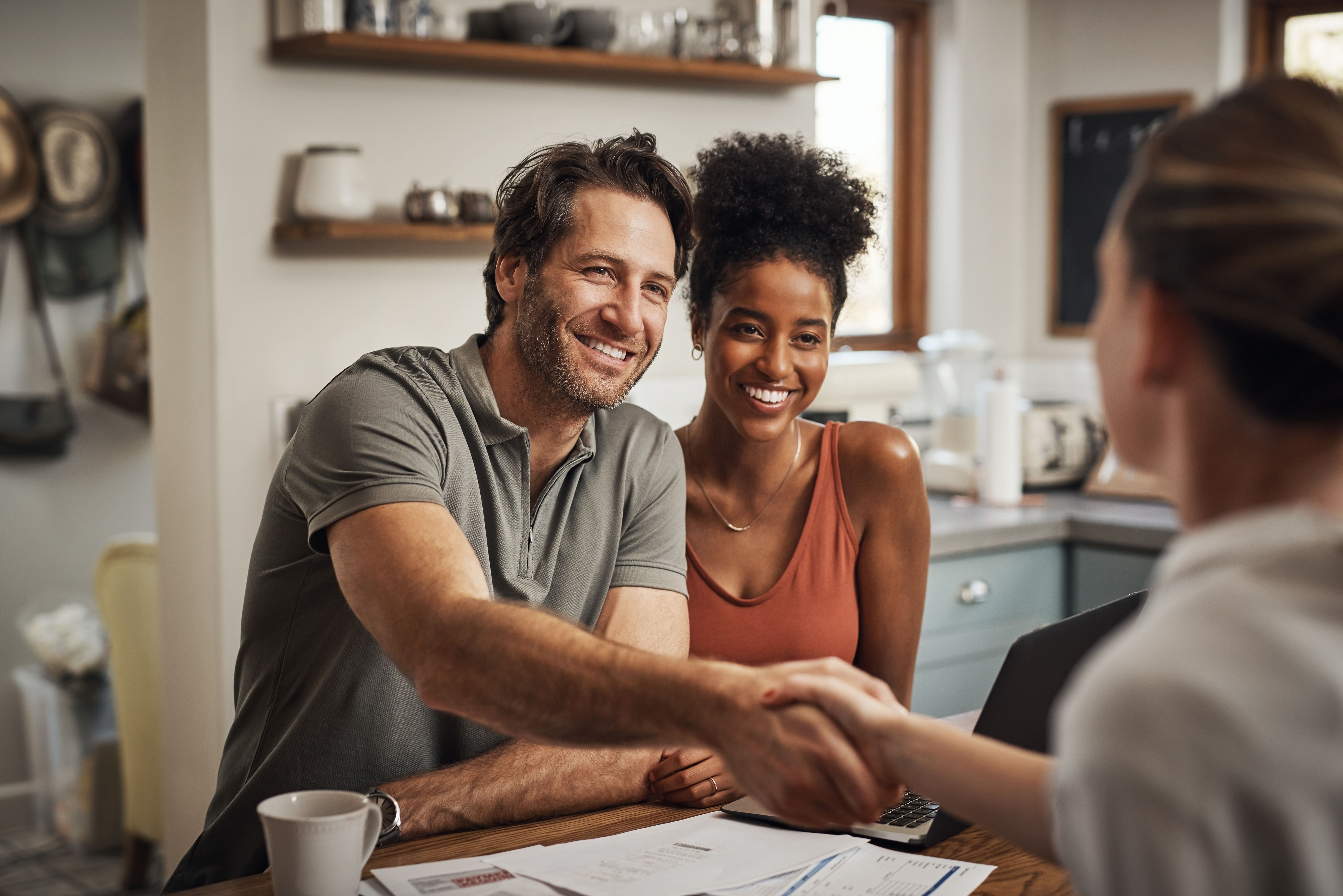 Smiling couple shaking hands with a professional over a desk with paperwork and a laptop in a home setting.
