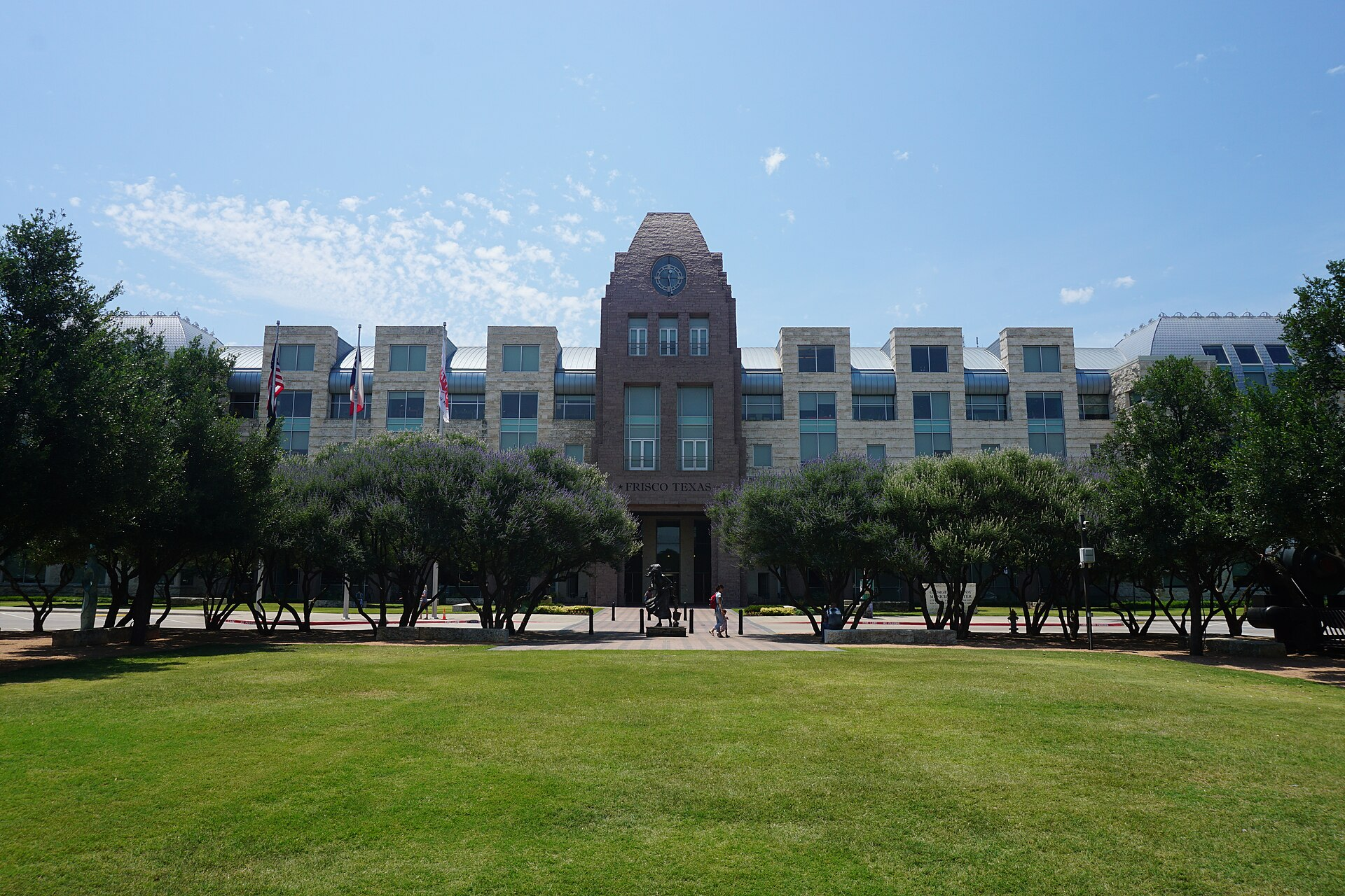 Front view of a large government building with a clock tower and multiple windows, labeled 'Frisco Texas,' surrounded by trees and a green lawn under a partly cloudy sky.