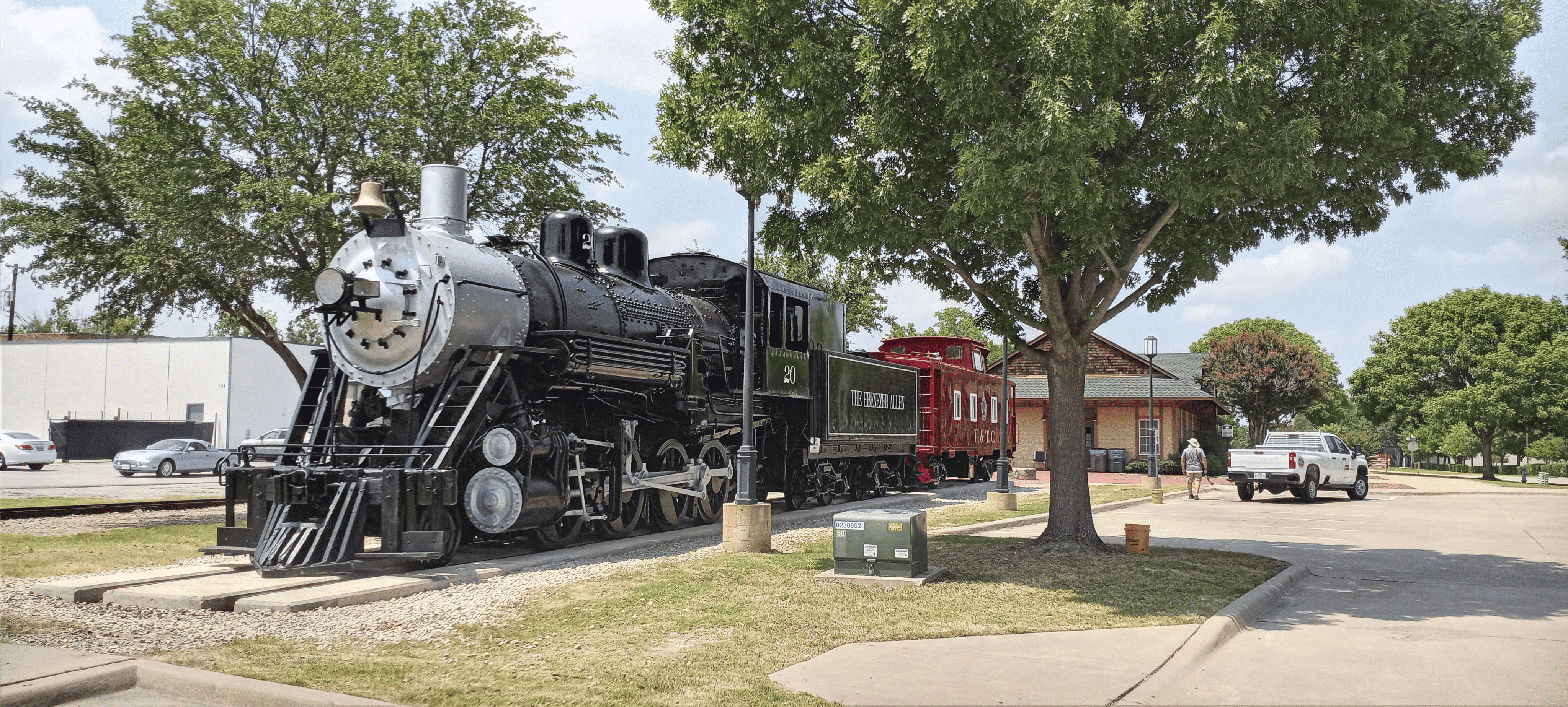 Historic black steam locomotive numbered 20 with a silver front and red caboose behind, displayed on short tracks beside a tree-lined sidewalk and a small building.