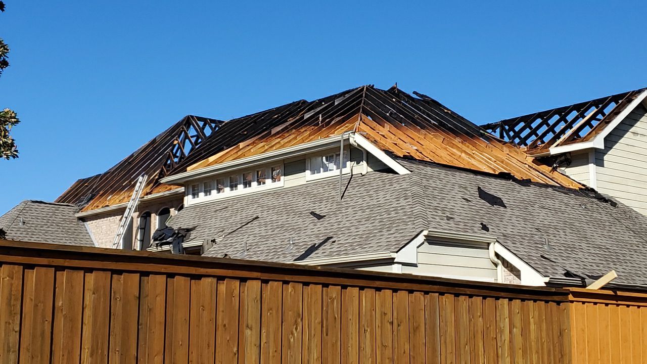 House roof severely damaged by fire, with charred and exposed wooden beams, against a clear blue sky, partially obstructed by a wooden fence.