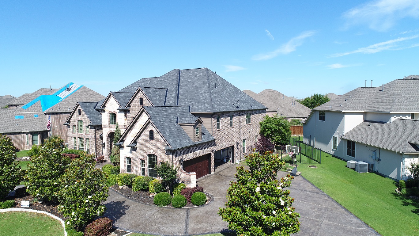 Large brick house with a dark gray shingled roof on a sunny day, surrounded by well-maintained green lawns, trees, and neighboring houses, one of which has a roof covered with blue tarp for repair.