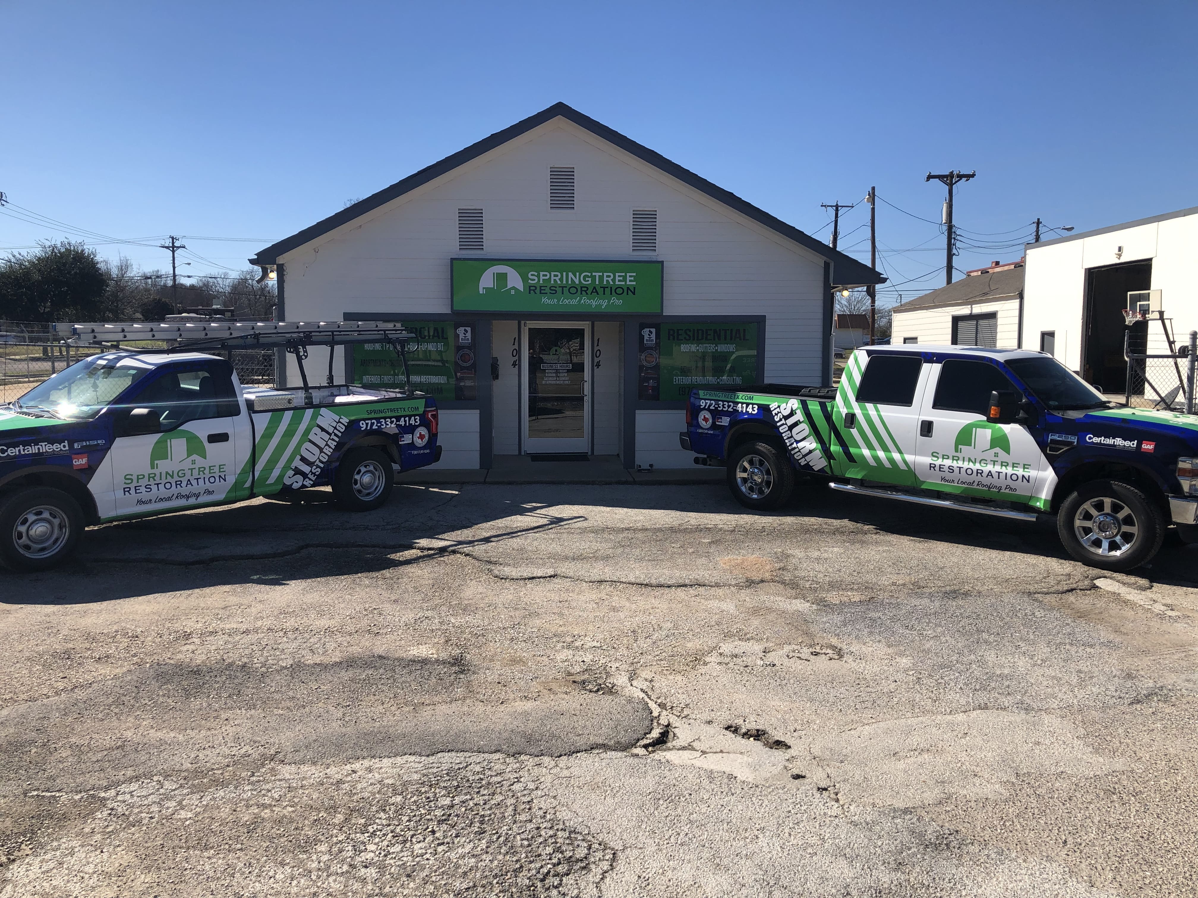 Two pickup trucks with SpringTree Restoration logos and green stripes parked in front of a white building with a green SpringTree Restoration sign above the entrance under clear blue sky.