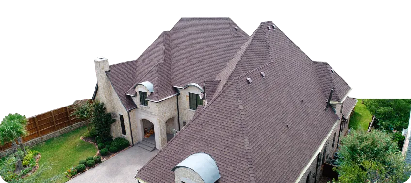 Aerial view of a large beige stone house with a complex brown shingle roof and surrounding green lawn with trees and shrubs.