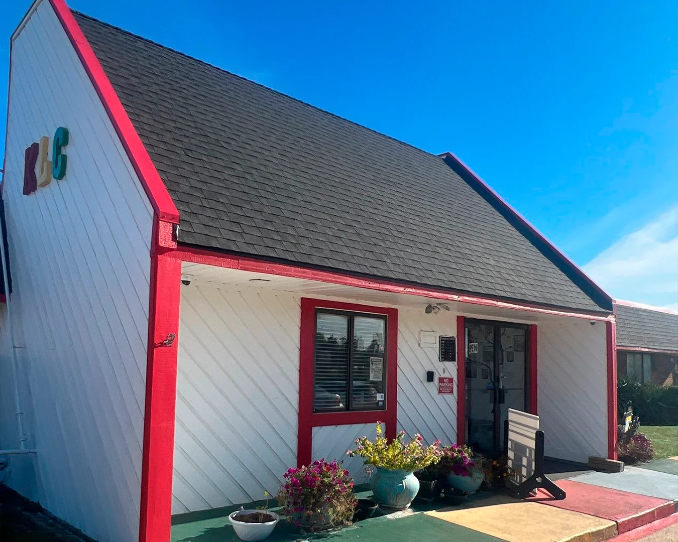 Small white building with red trim and a black roof under a clear blue sky, with flower pots near the entrance.