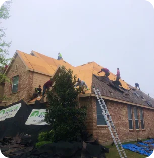 Several workers installing wooden sheathing on a house roof with a ladder leaning against the brick wall.